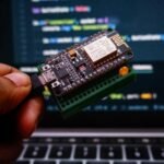 A close-up of a hand holding an Arduino microcontroller connected via USB, with blurred code displayed on a laptop screen in the background.