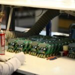 A technician inspecting multiple circuit boards laid out in a production line, with tools and cleaning spray nearby.