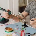 Technician soldering a circuit board with a soldering iron and magnifier, the job a fume extractor is for